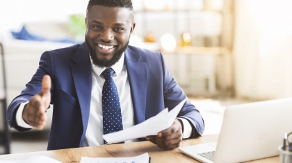 Partnership concept. Smiling young african american businessman holding documents and extending hand to shake, office interior, copy space